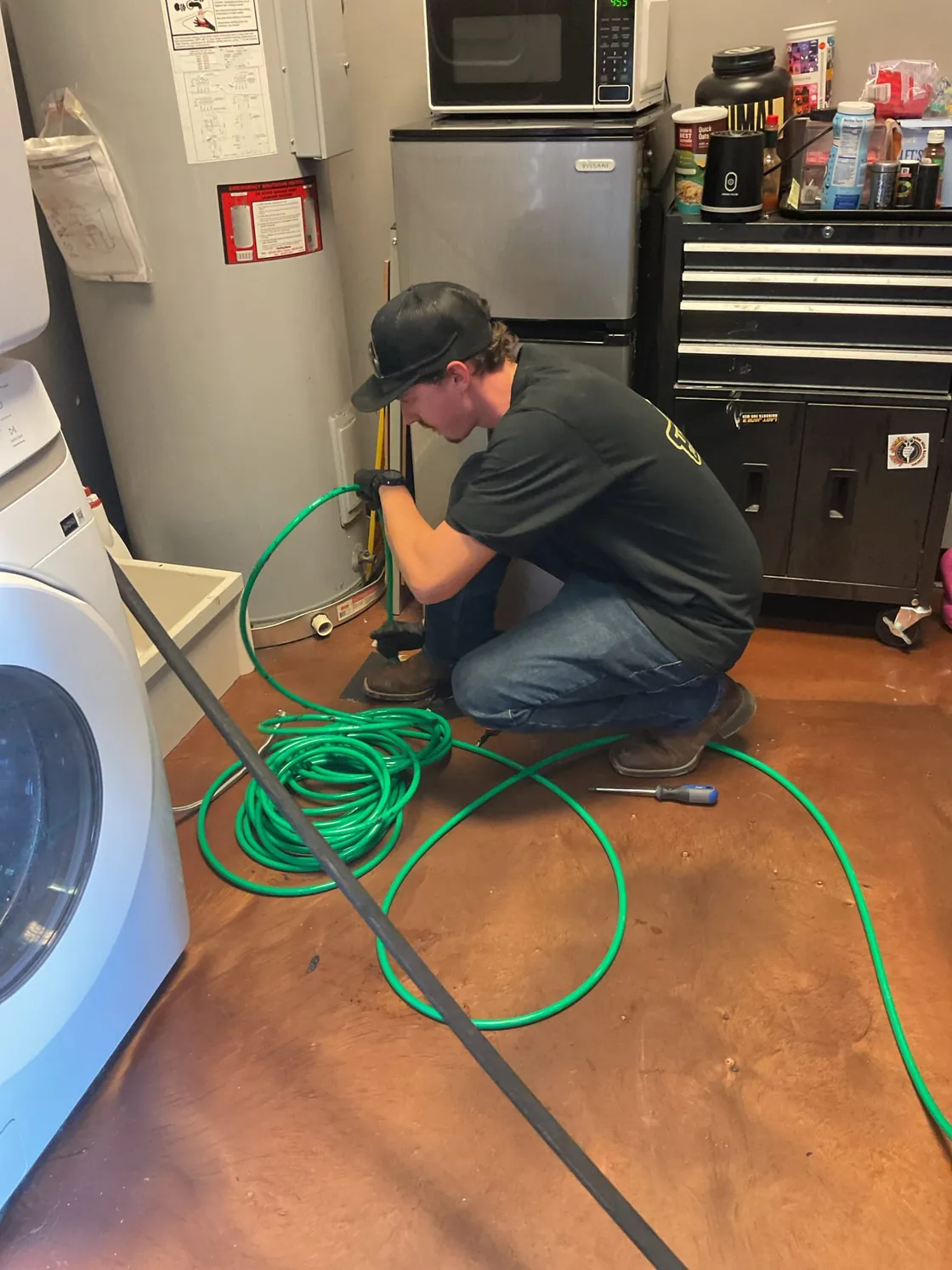 A technician using a green jetting hose beside a water heater