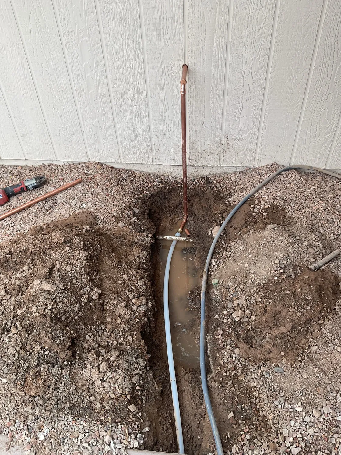 An excavated water line trench beside a home's exterior wall with new copper pipe and a gray supply line